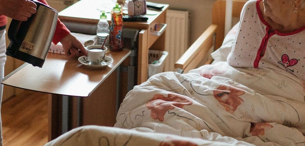 A caregiver serves tea to a senior woman in a cozy bedroom setting, fostering comfort and care.
