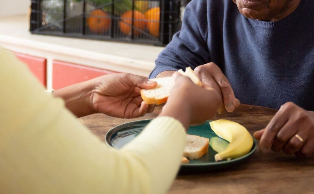 A woman assists an elderly man with meals at home, showcasing care and support.